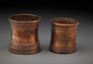 Two cylindrical copper containers with slightly flared rims and bases and horizontal bands of incised decorative patterns near the top and bottom.