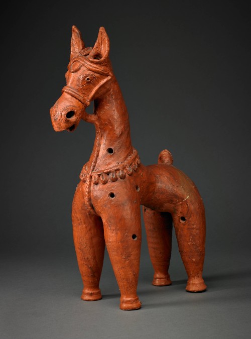 A stylized, reddish-brown terracotta figurine of a horse. It has an elongated neck and thick cylindrical legs with flared feet. The head features prominent ears, an open mouth, and a bridle.