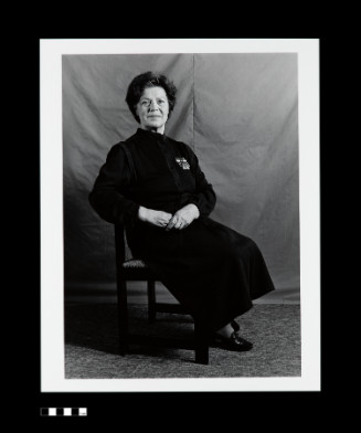 A portrait of a Soviet female sergeant. She is seated in front of a fabric background. She crosses her legs and clasps her hands while looking at the viewer.