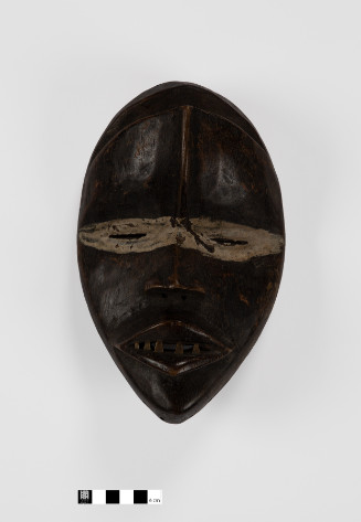 Oval-shaped wooden mask with inset metal teeth, central forehead ridge, and tan pigment around eyes.