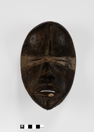Oval-shaped wooden mask with central forehead ridge, tan pigment around eyes, and an iron chin projection.