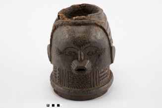 A dark wooden carving of a head with a hollow, rough-edged top. It features a circular forehead motif, incised lines by the eyes, semi-circular ears, a protruding open mouth, and a thick neck covered in carved geometric patterns. This view shows the front of one face.