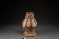 A painted cream-colored ceramic vessel with a flared base, bulbous body, and tapering neck. The center features alternating vertical red and black stripes. The neck, lower band, and base are detailed with intricate dark geometric, wavy, circular, and figurative patterns.