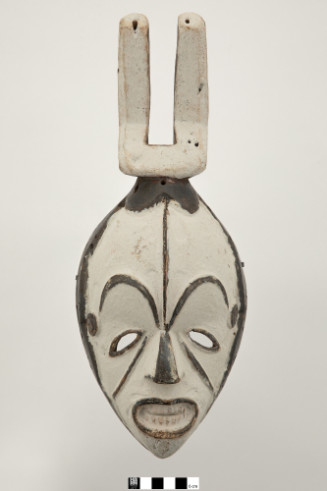 Elongated white wooden mask with arched eyebrows, dark outlines around the eyes, and an open mouth. A vertical prong-like crown structure extends from the top of the head.