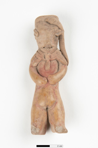 A flat tan clay figurine with a tall, ribbed headdress featuring a central red stripe. The face has slanted eyes, pierced circular cheeks, and a snout-like nose. Its small arms rest on its chest above long, separated legs.






