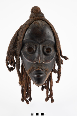Mask of stylized face with eye cylinders, braided hair, and a blackened surface.