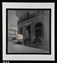 Water tank parked outside a corner arcade, while nearby, a man fine tunes a bicycle.