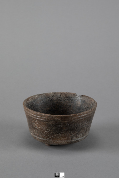 Small bowl with slightly rounded base supported on small tripod nubbins, area between nubbins is slightly concave.  Wall rises straight with slight outward slope to rim.  2 inscribed bands near rim, another near bottom frame panel area - 2 inscribed monkey figures on opposite sides - reclined with knees bent, hands held out in front, long tail curling out behind.