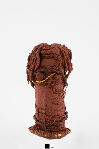 Cylindrical form wrapped with fiber, and hair and long braided strands on top. Glass or plastic bead strands on top and neck (yellow and blue beads) and wood or other brown beads (?) encircling form. Entire form is covered with red ochre